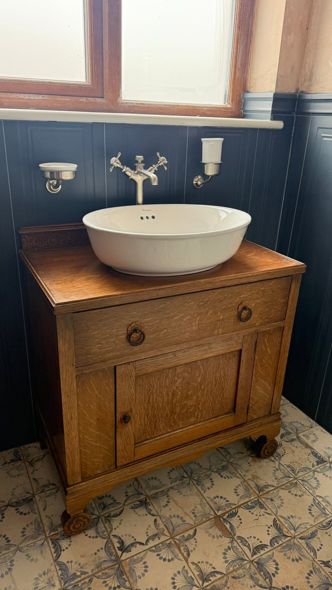 Antique wooden washstand with a round vessel basin and a wall-mounted bridge tap against navy panelling